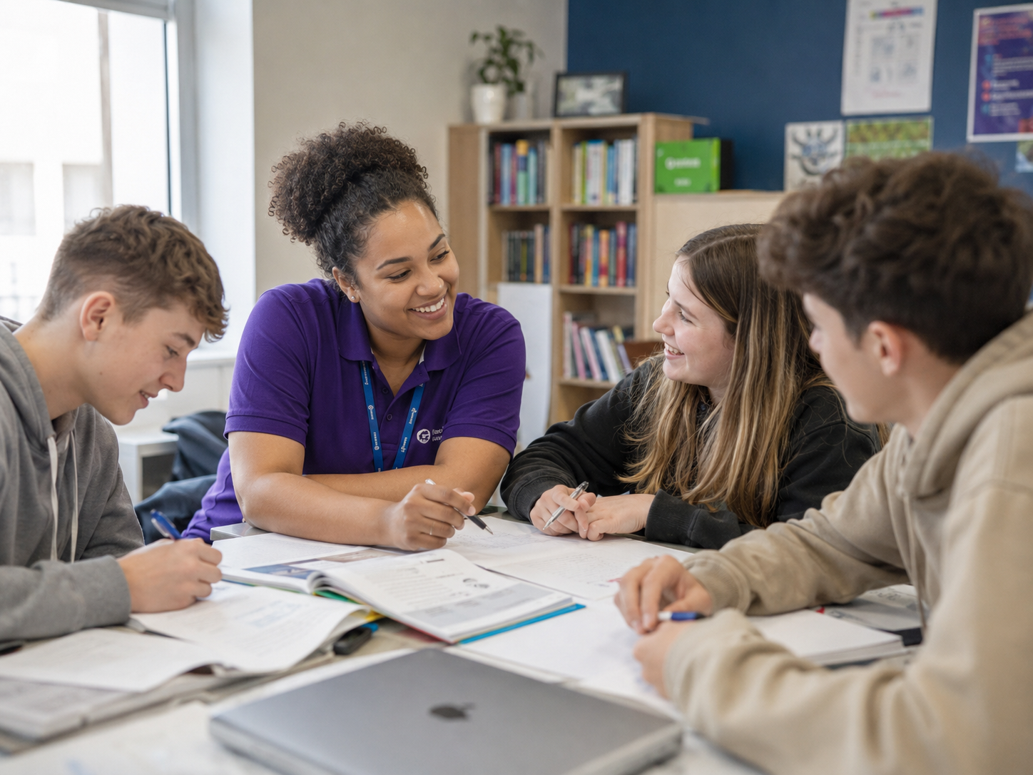 A care worker mentoring three teenagers at a study table with books and a laptop